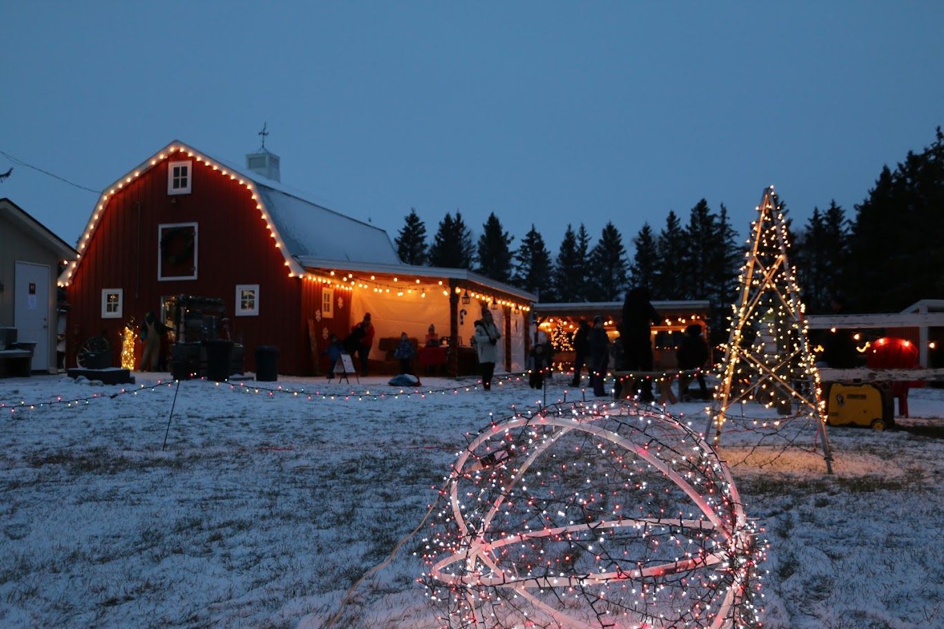The barn and buildings outlined in lights with a glowing light-wrapped tree and sphere in the snow